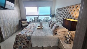 Ein Schlafzimmer mit einem großen Bett, einem flachbildfernseher an der Wand, einem Fenster mit Meerblick und einem Nachttisch mit Lampe.