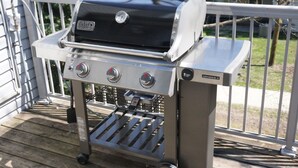 Ein Weber Genesis II Gasgrill mit drei Reglern und einem Seitenbrenner, montiert auf einem Metallgestell mit einer gerillten Ablage darunter.