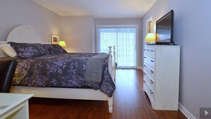 Ein Schlafzimmer mit einem Bett, einem Fernseher auf einer Kommode, einem Fenster mit Jalousien und einem Nachttisch mit Lampe.