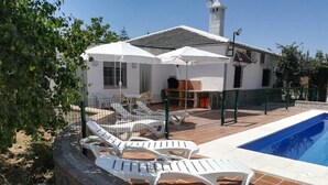 Un espace aménagé au bord de la piscine, avec des chaises longues et des parasols, une terrasse carrelée et un bâtiment doté d’une cheminée.