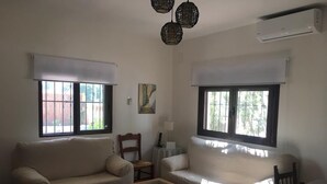 Un salon avec deux canapés, une table basse et une table à manger. Il y a des fenêtres avec des stores, un tableau au mur et un ventilateur de plafond.