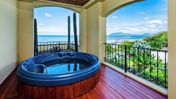 Un balcón con jacuzzi, suelo de madera y vistas al océano y la vegetación.