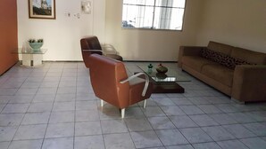 Ein Wohnzimmer mit einem braunen Sofa, einem Glastisch und einem Sessel. Es gibt ein Fenster mit Gitterstäben und ein Bild an der Wand.