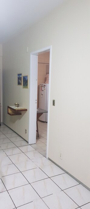 A hallway with a tiled floor, a wooden shelf with a vase, and a door leading to a room with a refrigerator.