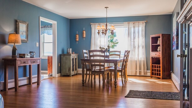 A dining area with a wooden table and chairs, a chandelier, and a view of the outdoors through a window with sheer curtains. - Located Directly On Rockland Harbor (Rockland)