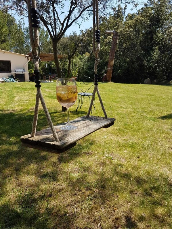 A rustic wooden swing suspended by ropes, with a glass of wine on it, set in a grassy outdoor area with trees and a building in the background.