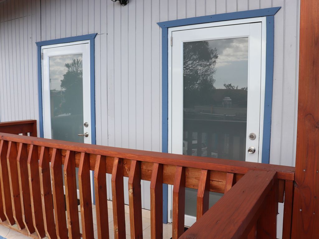 Eine Holzterrasse mit Geländer, zwei Türen mit blauer Umrandung und ein Fenster mit Glas, das Bäume und ein Gebäude reflektiert.