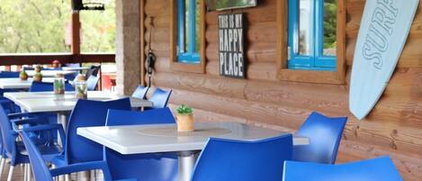 A wooden cabin with blue chairs and tables, a surfboard sign, and a TV on the wall.