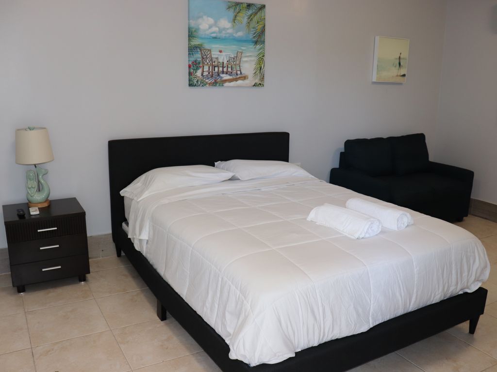 A neatly made bed with white bedding, a black headboard, and a black sofa in a room with a tiled floor.