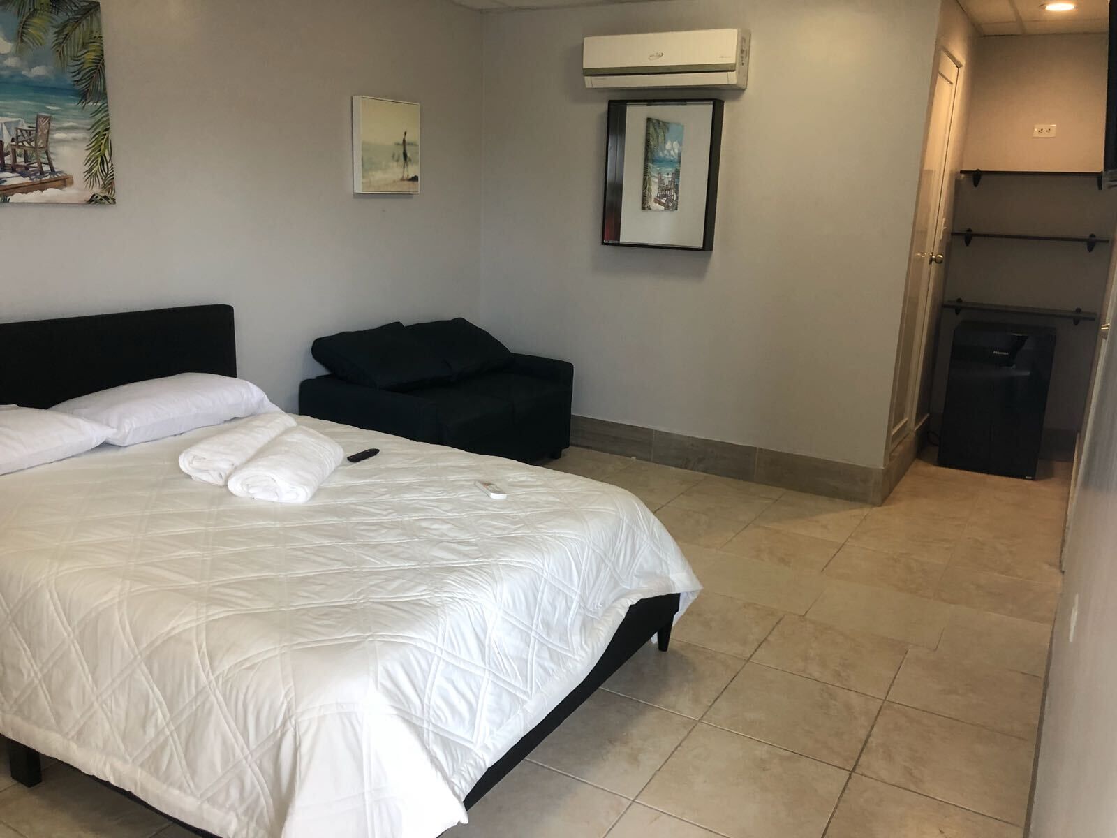 A hotel room with a bed, a black sofa, an air conditioning unit, and a small trash bin.