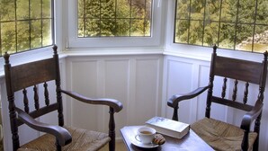 A corner room with two wooden chairs, a small table, and a view of trees through a window with a grid pattern.