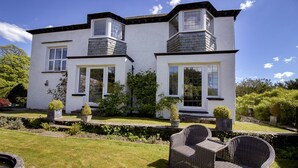 A two-story house with a white exterior, a stone chimney, and a garden with a wicker chair and potted plants.
