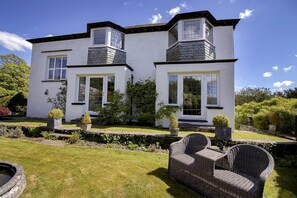 A two-story house with a white exterior, large windows, and a garden with a pond and outdoor furniture. - Beechmount Country House (Near Sawrey)