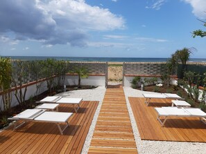 Une terrasse en bois avec des chaises longues blanches, un chemin de gravier et une vue sur la plage et l’océan.