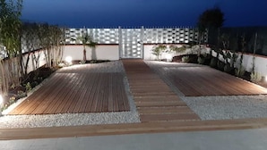 A well-lit outdoor area with a wooden deck, gravel pathways, and a white wall with a checkered pattern. - DORIDE Apartment on the beach (Balestrate)