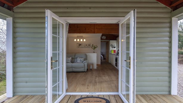 A wooden cabin with a welcoming mat, a sofa, and a kitchen area visible through open doors. - Peaceful, relaxing  lodge for 2 set in countryside (Elgin)