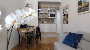 Un salon avec un canapé et un coin repas comprenant des chaises et une table. Au fond, on aperçoit une cuisine avec des placards et un four à micro-ondes.