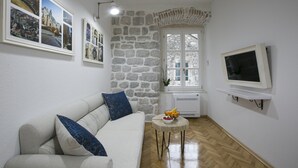 Un salon avec un mur en pierre, un canapé blanc, une petite table ronde sur laquelle on a disposé des fruits, et un téléviseur fixé au mur.