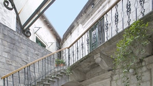 Un escalier en pierre menant à un balcon orné d’une balustrade en fer forgé, une porte verte et un pot de fleurs.