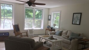 A living room with a sofa, coffee table, and a ceiling fan. There are large windows with a view of trees. - Pond House - Donnell's Pond in Franklin, Maine (Franklin)