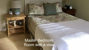 A master bedroom with a view, featuring a large bed, a nightstand with a lamp, a bedside table with a clock, and a wall-mounted telephone. - Pond House - Donnell's Pond in Franklin, Maine (Franklin)
