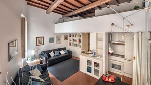 Ein moderner Wohnraum mit Schlafgalerie, einem schwarzen Sofa, einem Esstisch mit roter Ablage und einer Küche mit weißen Schränken und einem Herd.