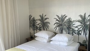A neatly made bed with white linens and a green throw blanket, a nightstand with a lamp, and a wall mural of palm trees. - Spacious T3 with large terrace SEA VIEW (Le Grau-du-Roi)