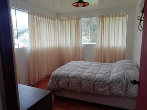 A bedroom with a bed, two windows with curtains, a wooden floor, and a ceiling light fixture. - Hacienda Cachimayo, Cusco (Cachimayo)
