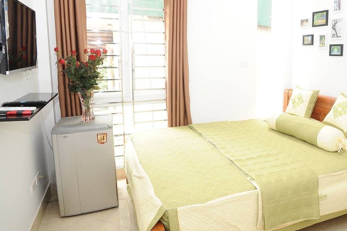 A bedroom with a bed, a small desk, a refrigerator, a TV, and a wooden shelf.