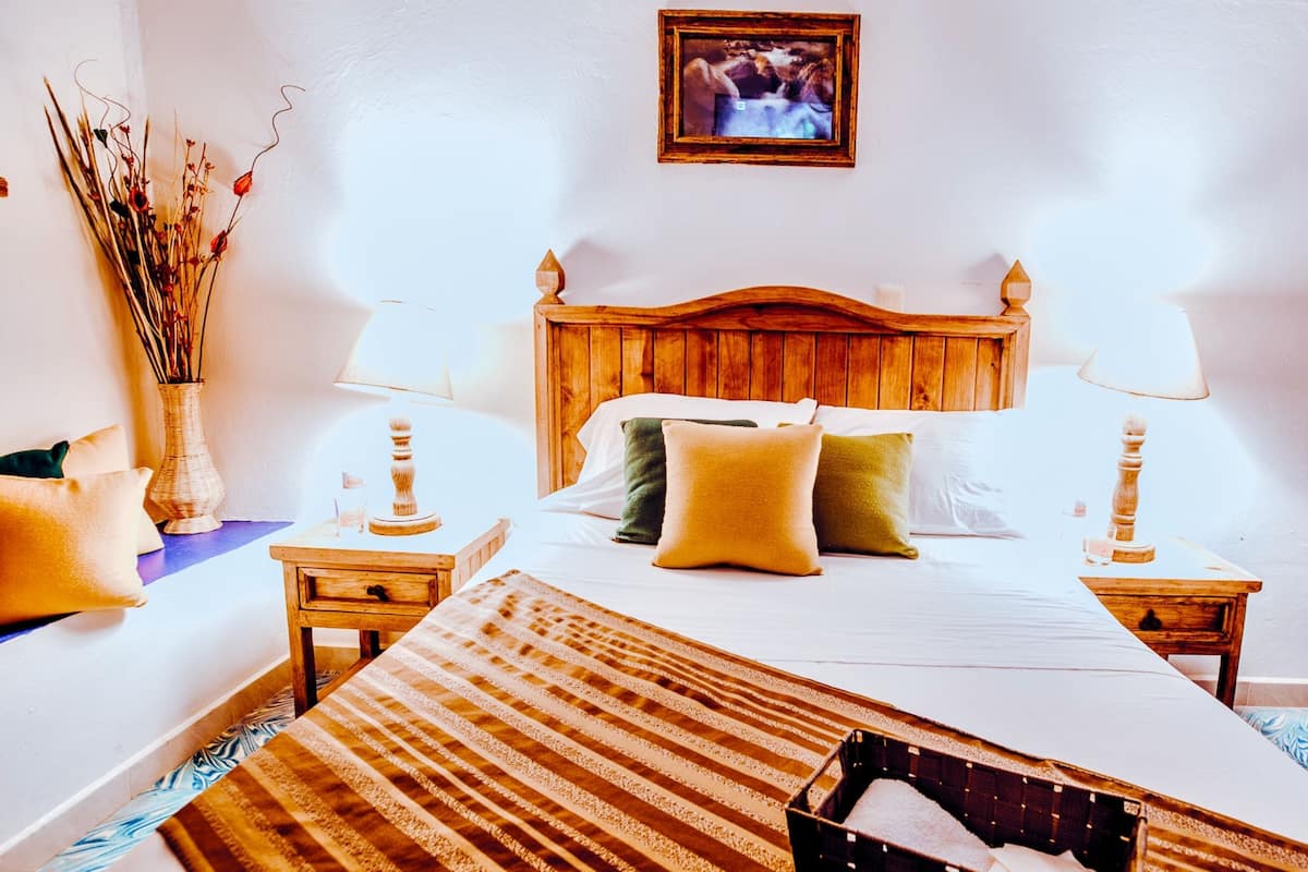 Ein ordentlich eingerichtetes Schlafzimmer mit einem hölzernen Kopfteil, einem Bett mit weißen und farbigen Kissen, zwei Nachttischen mit Lampen, einer Vase mit Trockenblumen und einem gerahmten Bild an der Wand.