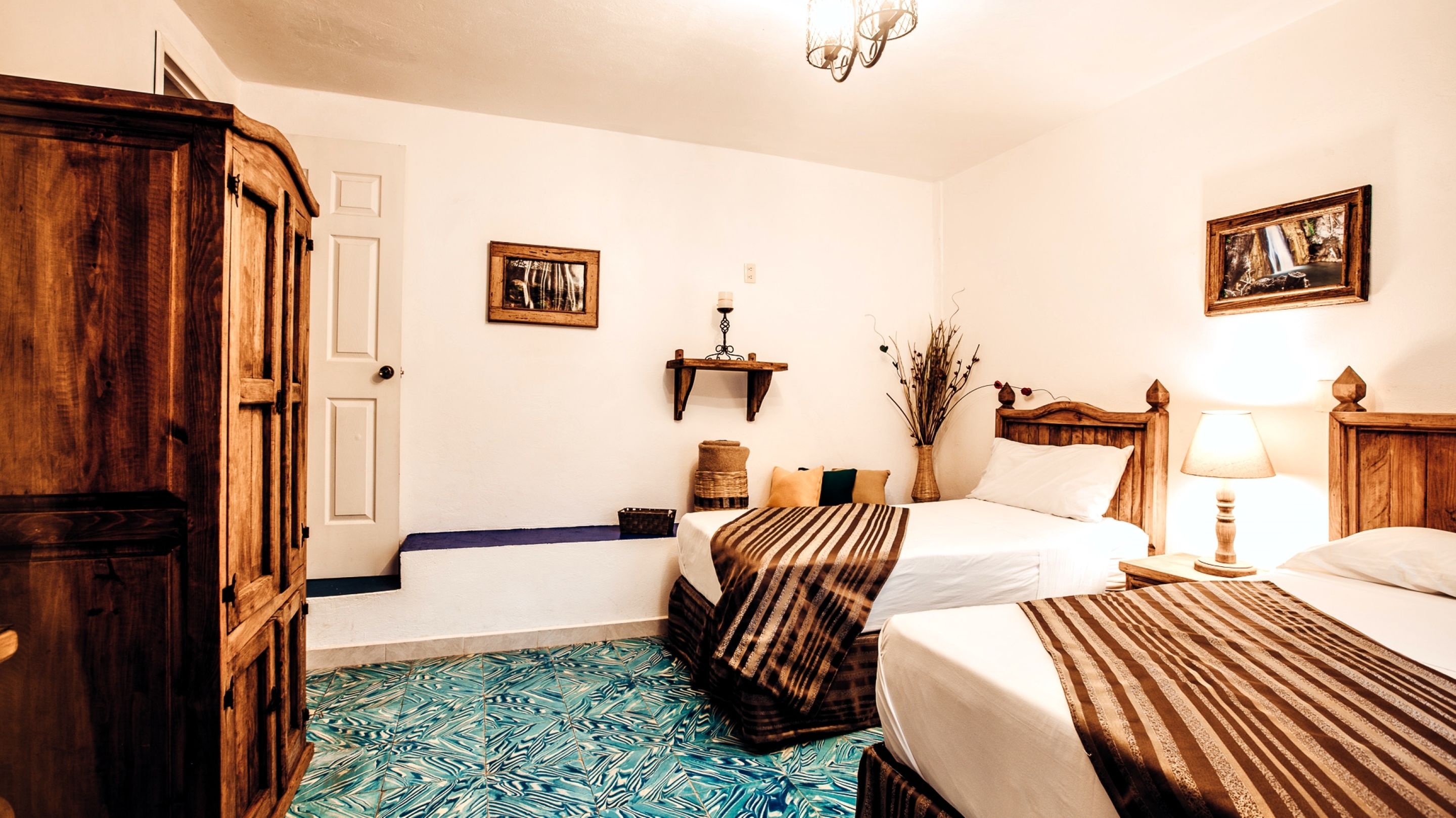 Une chambre d’hôtel avec deux lits, une armoire en bois, un sol à motifs, une étagère fixée au mur avec une bougie, et un tableau encadré.
