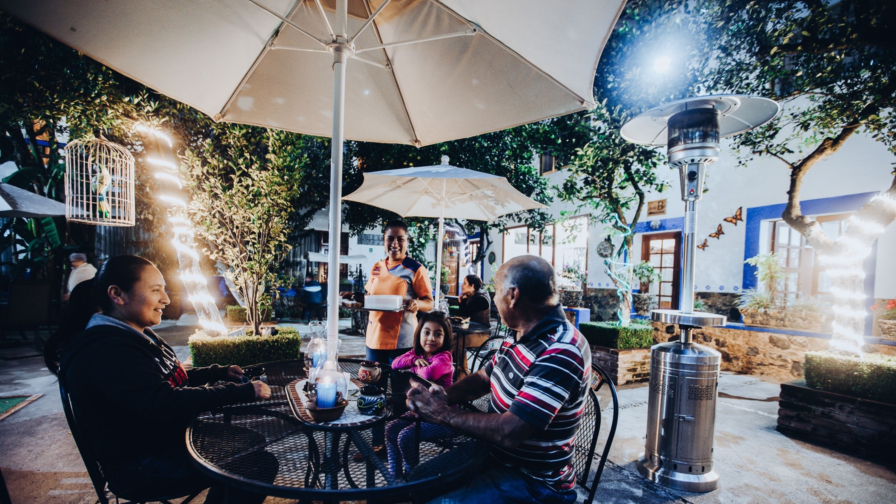 Un área de comedor al aire libre con mesas y sillas, rodeada de sombrillas y luces decorativas.