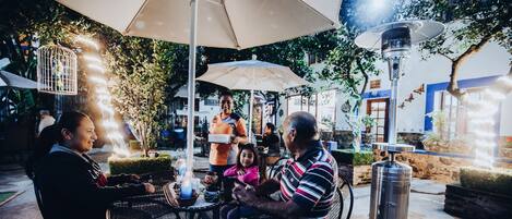 Un área de comedor al aire libre con mesas y sillas, rodeada de sombrillas y luces decorativas.