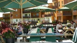 Un área de comedor al aire libre con mesas y sillas bajo sombrillas verdes, rodeada de edificios de madera con balcones.
