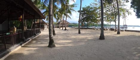 Une plage de sable bordée de palmiers et surmontée d’une structure en bois, probablement un abri de plage ou un restaurant, avec une vue sur la mer en arrière-plan.