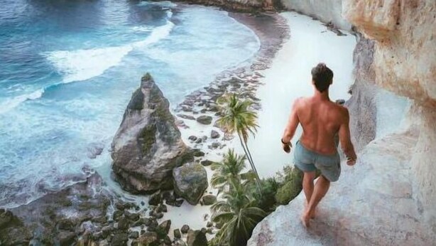 Un hombre camina por una costa rocosa con aguas azul cristalino y una palmera.