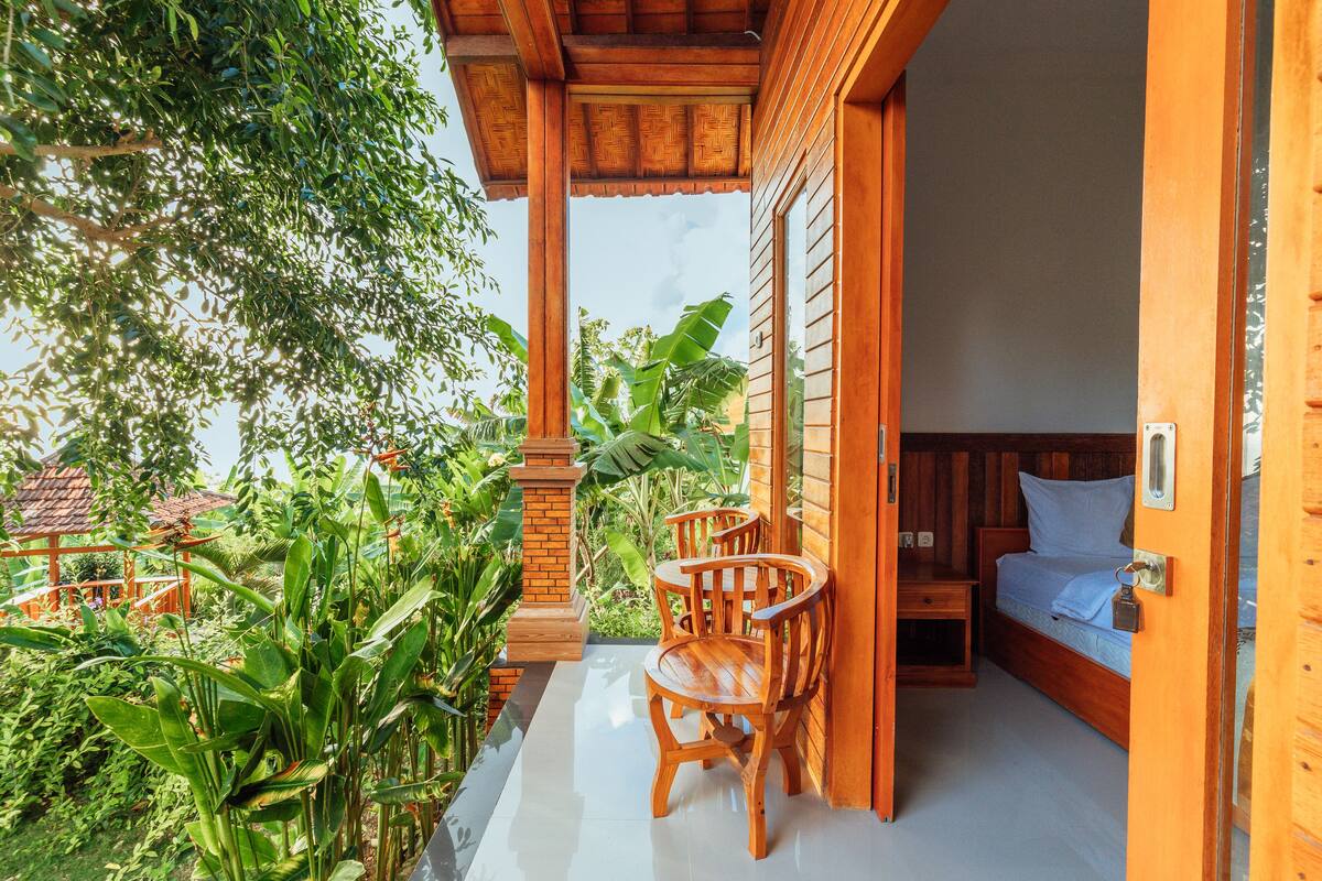 A wooden balcony with a bed and two chairs, surrounded by lush greenery.