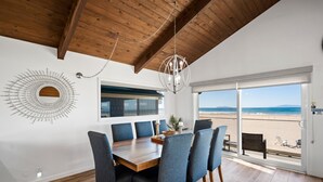 Comedor con mesa de madera, sillas azules, una lámpara de araña y vista a la playa a través de amplios ventanales.