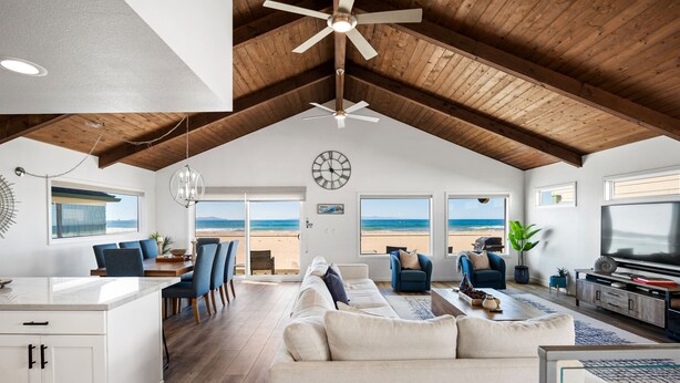 Un amplio salón con techo alto, vigas de madera, un televisor grande y vista a la playa.