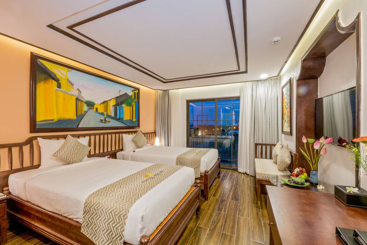 A hotel room with two beds, a balcony with a view, a desk with a vase of flowers, and a painting on the wall.