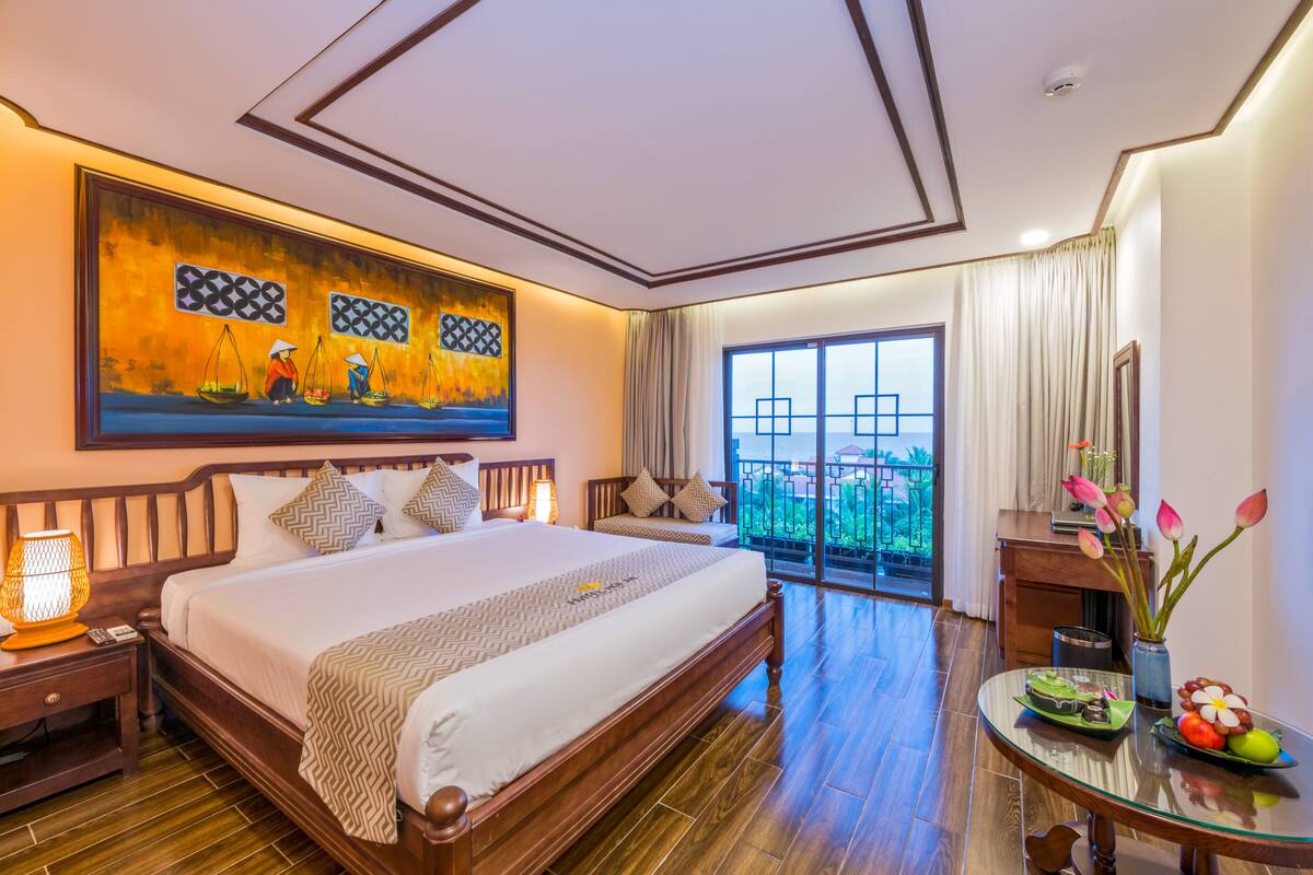 A hotel room with a large bed, a painting on the wall, a balcony with curtains, and a table with a vase of flowers.