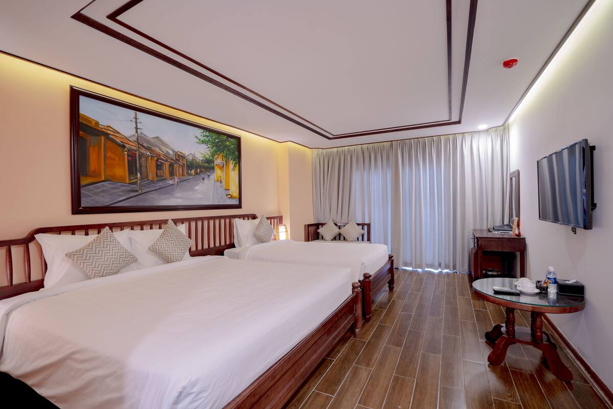 A hotel room with two beds, a painting of a street scene, a TV, and a small table with a bottle.