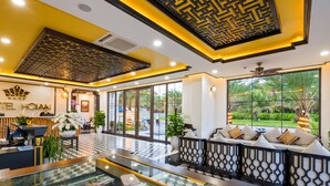 A hotel lobby with a reception desk, seating area, and large windows offering a view of the outdoors. - Fivitel Hoi An Hotel (Da Nang)