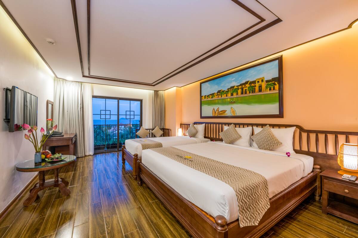 A hotel room with two large beds, a painting on the wall, a table with flowers, and a view of the outdoors through a window.