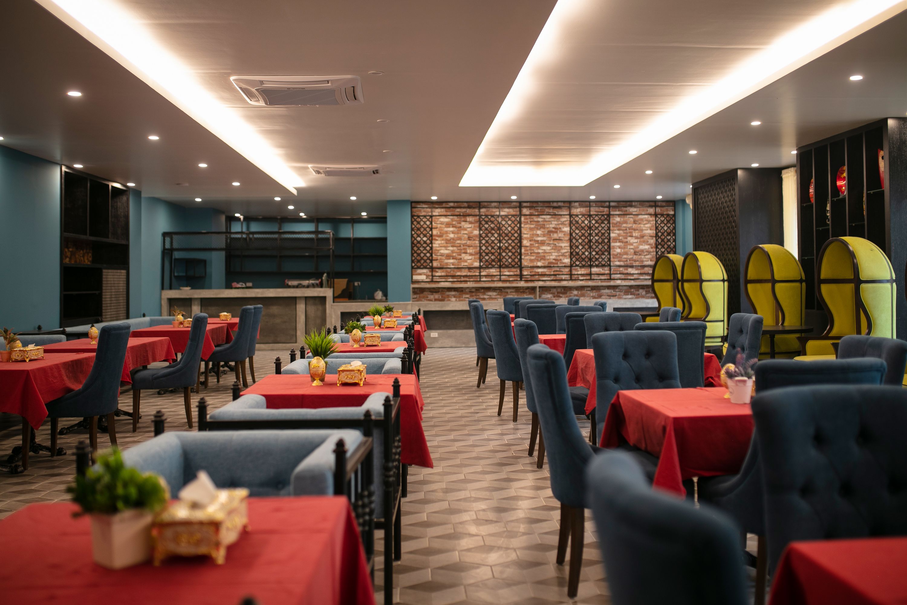 Un restaurante con mesas dispuestas, manteles rojos y sillas azules, una pared de ladrillo con un panel decorativo y un techo con iluminación empotrada.