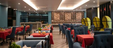 Un restaurante con mesas dispuestas, manteles rojos y sillas azules, una pared de ladrillo con un panel decorativo y un techo con iluminación empotrada.