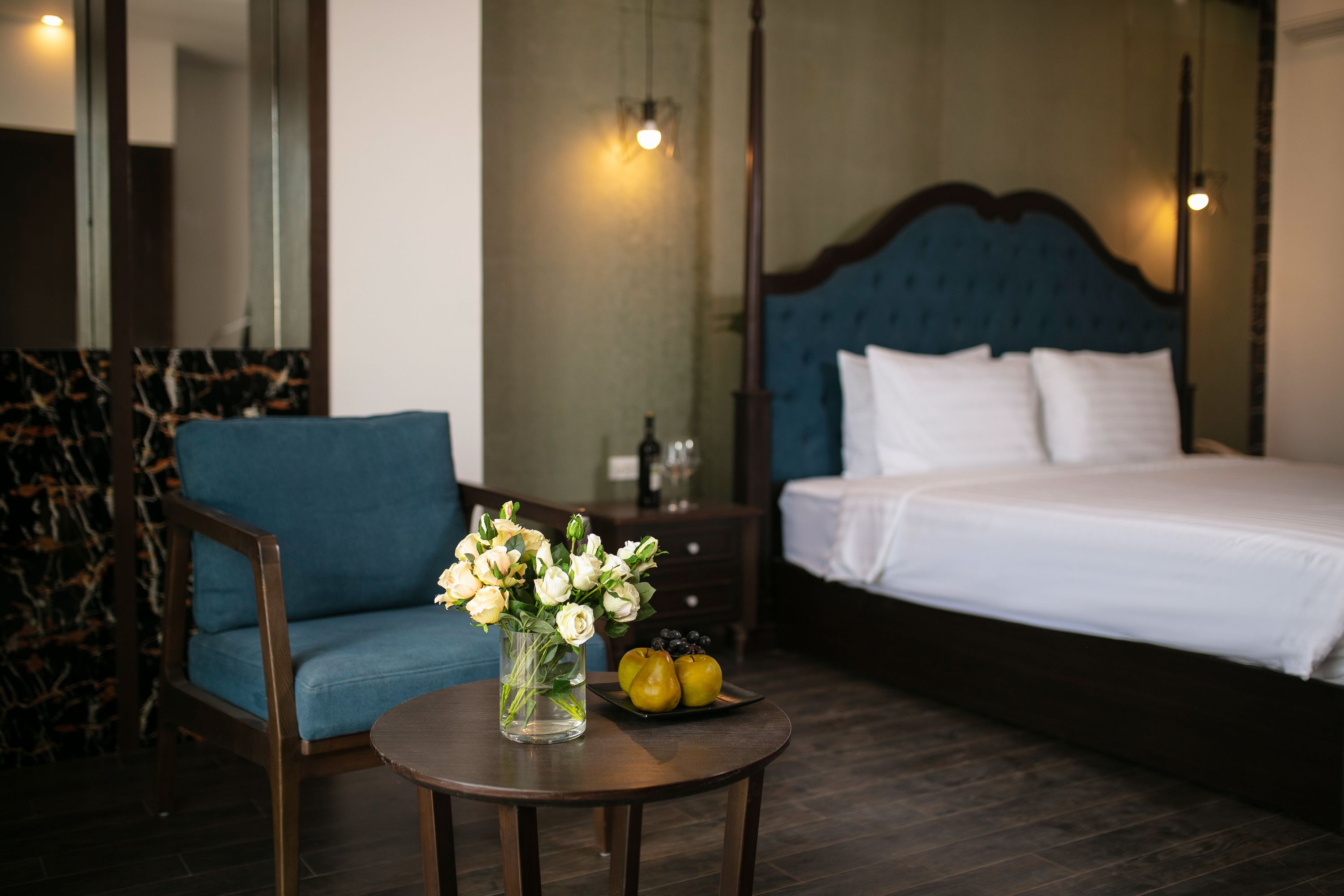 Habitación de hotel con cama, una silla, una mesa con un florero y una bandeja de frutas.