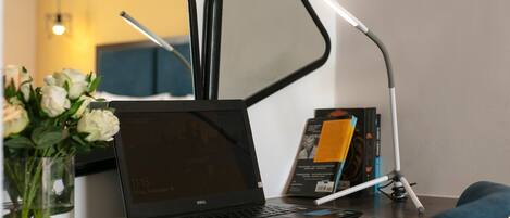 A hotel desk with a laptop, a mirror, a lamp, and a vase with flowers.