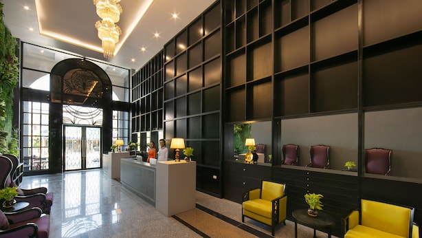 A lobby with a reception desk, seating area with yellow chairs, and a large wall with shelves. - Azalea Parkview Hotel (Vientiane)