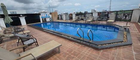Una piscina en la azotea con solárium empedrado, sillas de descanso y vistas al horizonte de la ciudad.